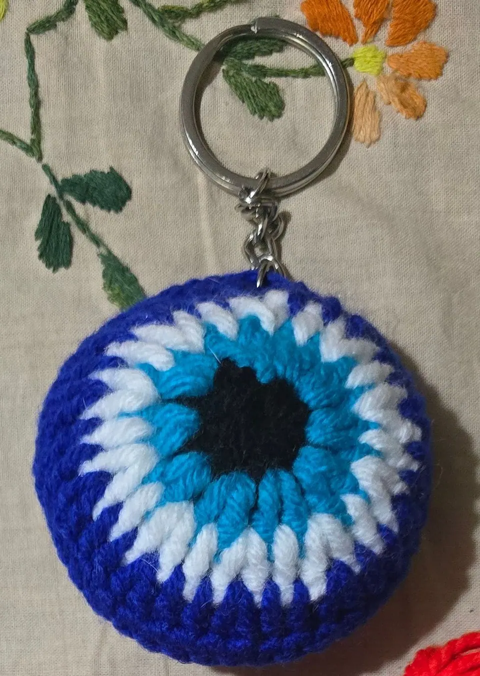 Round crochet evil-eye keychain in royal blue, white and sky blue with a black centre, on a metal key ring