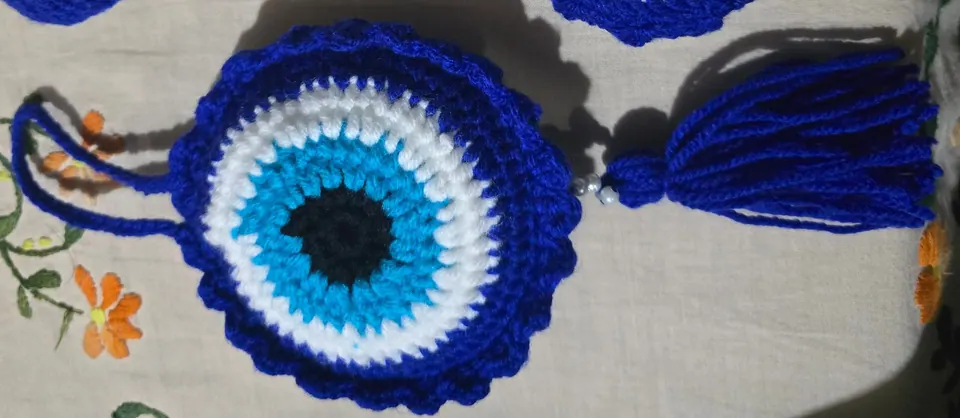 Large crochet evil-eye charm lying flat with its yarn loop on one side and blue tassel on the other