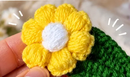 Yellow crochet flower hair clip with a white centre and green leaf, held between fingers (illustrative image)