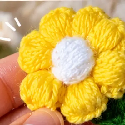Yellow crochet flower hair clip with a white centre and green leaf, held between fingers (illustrative image)