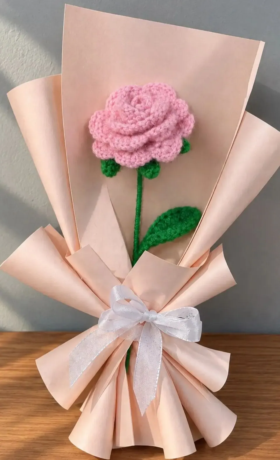 Pink crochet rose bouquet wrapped in blush paper with a white ribbon bow, standing on a wooden table